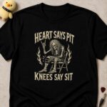 Black t-shirt printed HEART SAYS PIT above and KNEES SAY SIT below a skeleton throwing devil horns from a folding chair