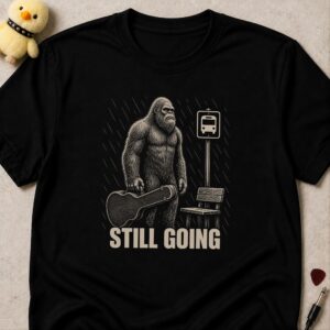 Black t-shirt printed STILL GOING below bigfoot carrying a guitar case at a bus stop in the rain