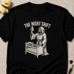 Black t-shirt printed THE NIGHT SHIFT above a hooded skeleton reaper holding a baby bottle beside a cot