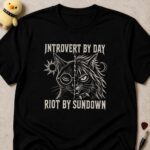 Black t-shirt printed INTROVERT BY DAY RIOT BY SUNDOWN around a cat's face split between a calm half and a snarling half