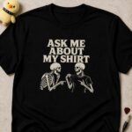 Black t-shirt printed ASK ME ABOUT MY SHIRT above two skeletons, one pointing at the other's shirt