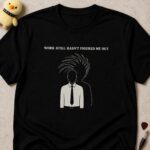 Black t-shirt printed WORK STILL HASN'T FIGURED ME OUT above a man in a shirt and tie whose shadow has a mohawk